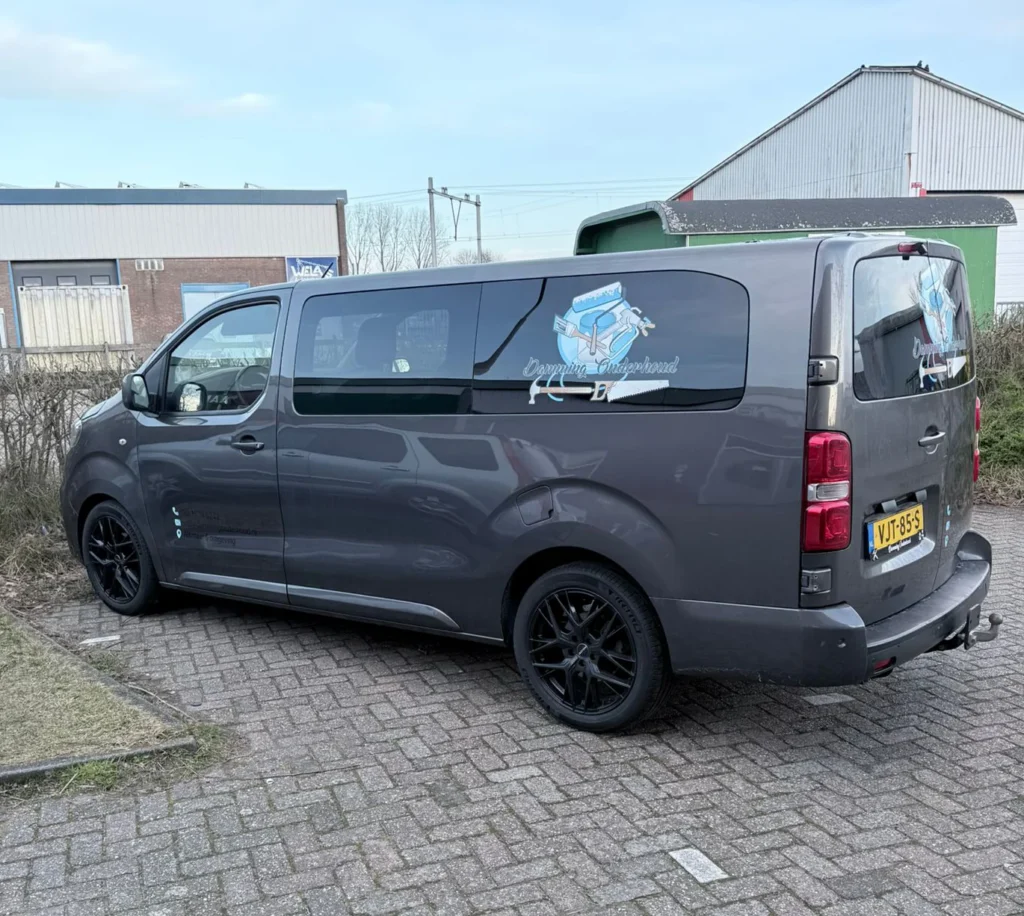 Donkergrijze bedrijfsbus met logobelettering op de zijruit, geparkeerd op een klinkerterrein in Castricum